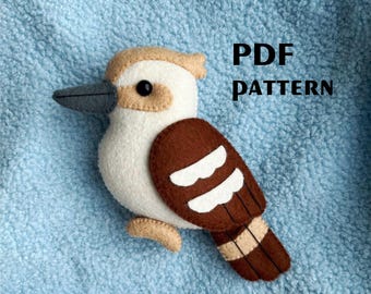 May include: A felt kookaburra bird with a white body, brown wings, and a beige head. The bird has a black eye and a gray beak. The text "PDF PATTERN" is displayed in black.