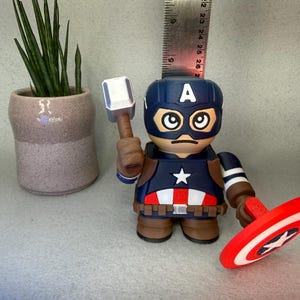 Op de afbeelding: Een Captain America-beeldje met een blauwe helm en de letter "A", met een hamer en schild. Het beeldje heeft een rood, wit en blauw kleurenschema. Op de achtergrond staat een kleine potplant.