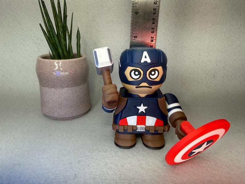 Op de afbeelding: Een Captain America-beeldje met een blauwe helm en de letter "A", met een hamer en schild. Het beeldje heeft een rood, wit en blauw kleurenschema. Op de achtergrond staat een kleine potplant.