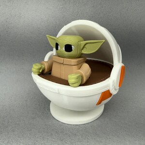 May include: A white and orange plastic toy spaceship with a green and brown figure sitting inside. The figure has large ears and big eyes.