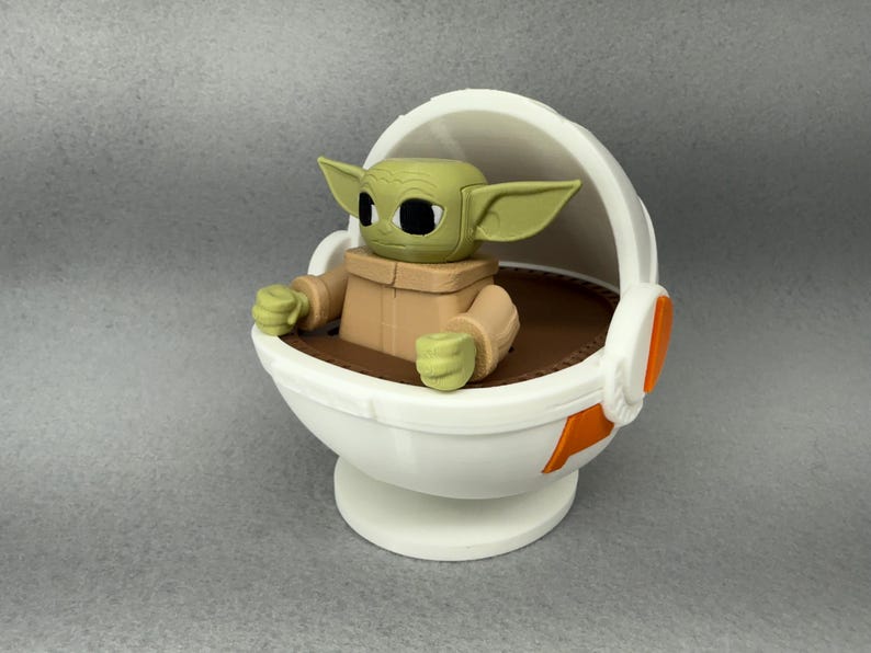 May include: A white and orange plastic toy spaceship with a green and brown figure sitting inside. The figure has large ears and big eyes.