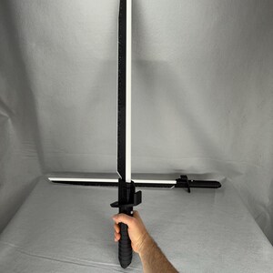 May include: A black and white sword with a crossguard and a textured handle. The blade is white with a black edge and a black spine. The sword is held in a hand.