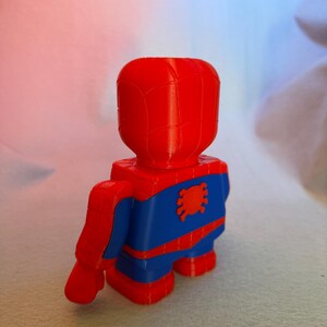 May include: A red and blue 3D printed figurine of Spider-Man. The figurine is standing with its back to the camera. The figure has a red head and torso, blue legs, and a red spider symbol on its chest.
