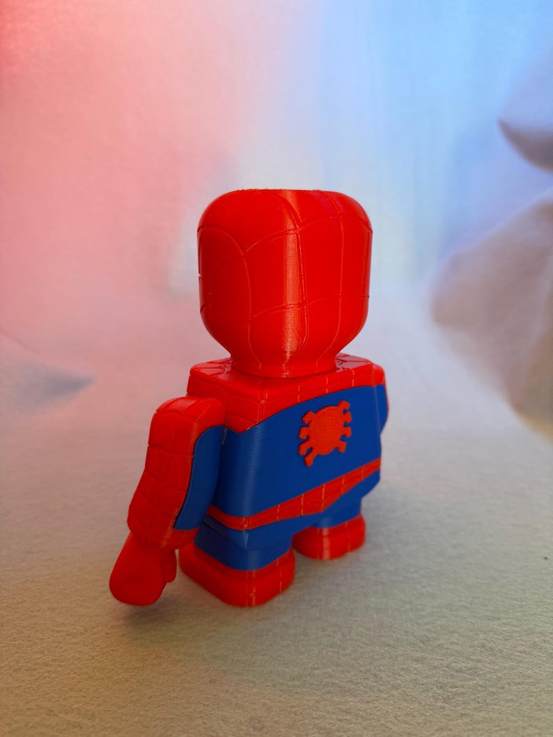 May include: A red and blue 3D printed figurine of Spider-Man. The figurine is standing with its back to the camera. The figure has a red head and torso, blue legs, and a red spider symbol on its chest.