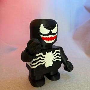 May include: Black and white 3D printed figurine of Venom, a Marvel Comics superhero. The figurine is in a standing pose with its arms outstretched. The figurine has a black body with white eyes, teeth, and a spider symbol on its chest.