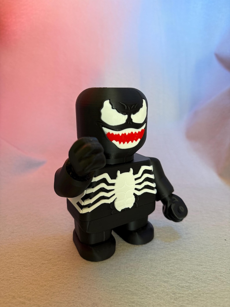 May include: Black and white 3D printed figurine of Venom, a Marvel Comics superhero. The figurine is in a standing pose with its arms outstretched. The figurine has a black body with white eyes, teeth, and a spider symbol on its chest.