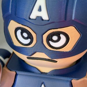Op de afbeelding: Een close-up van een Captain America-beeldje. Het beeldje heeft een blauwe helm met een witte "A" op het voorhoofd. Het gezicht is bruin met zwart omlijnde ogen en een zwarte lijn voor een mond. De borst heeft een witte ster.