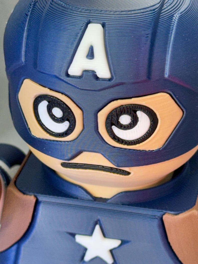 Op de afbeelding: Een close-up van een Captain America-beeldje. Het beeldje heeft een blauwe helm met een witte "A" op het voorhoofd. Het gezicht is bruin met zwart omlijnde ogen en een zwarte lijn voor een mond. De borst heeft een witte ster.