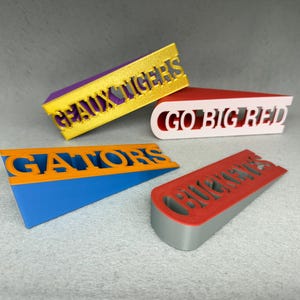 May include: Four colorful doorstops with different college team names. The doorstops are made of plastic and have a wedge shape. The names on the doorstops are "Geaux Tigers", "Go Big Red", "Gators", and "Buckeyes".
