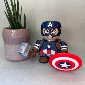Op de afbeelding: Een Captain America-beeldje met een blauwe helm en schild, met een hamer. Het beeldje heeft een ster op de borst en een rood, wit en blauw schild. Een kleine potplant staat op de achtergrond.