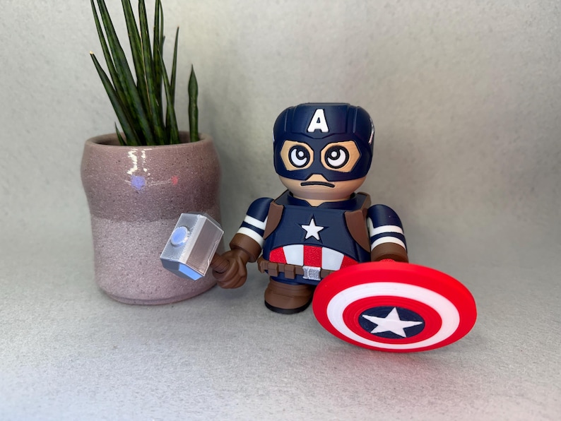 Op de afbeelding: Een Captain America-beeldje met een blauwe helm en schild, met een hamer. Het beeldje heeft een ster op de borst en een rood, wit en blauw schild. Een kleine potplant staat op de achtergrond.