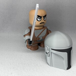 May include: A gray and black Mandalorian helmet with a white lightsaber held by a brown and gray action figure with a stern expression.
