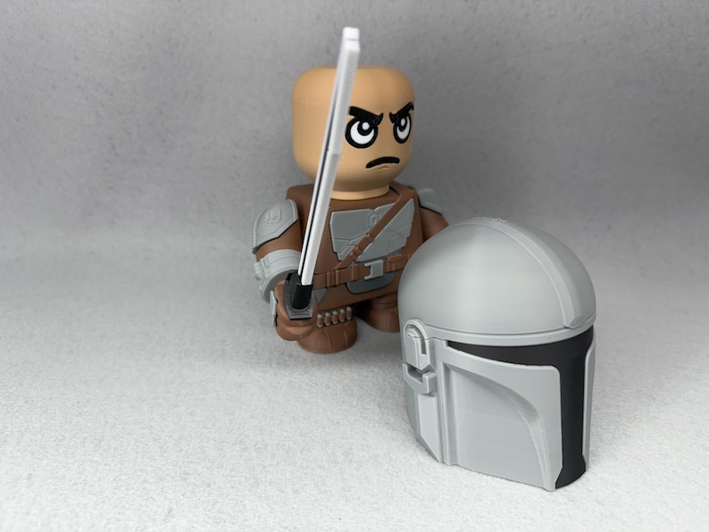 May include: A gray and black Mandalorian helmet with a white lightsaber held by a brown and gray action figure with a stern expression.