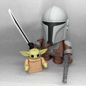 May include: A gray and brown Mandalorian action figure holding a black blaster rifle and a white lightsaber. A small green and brown Grogu action figure stands next to him.