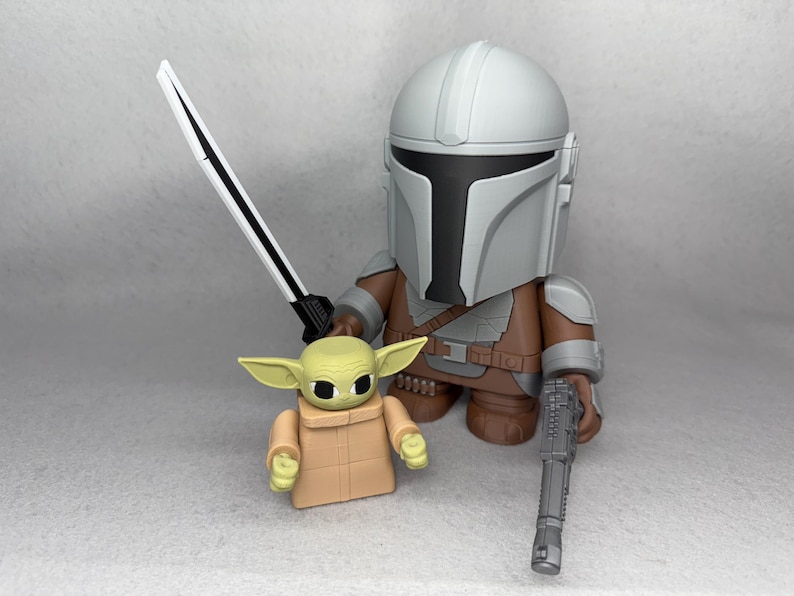May include: A gray and brown Mandalorian action figure holding a black blaster rifle and a white lightsaber. A small green and brown Grogu action figure stands next to him.