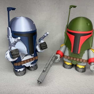 May include: Two Star Wars action figures, one in blue and silver and the other in green and red, stand on a gray surface. The blue and silver figure is holding a blaster rifle. The green and red figure is holding a blaster pistol.