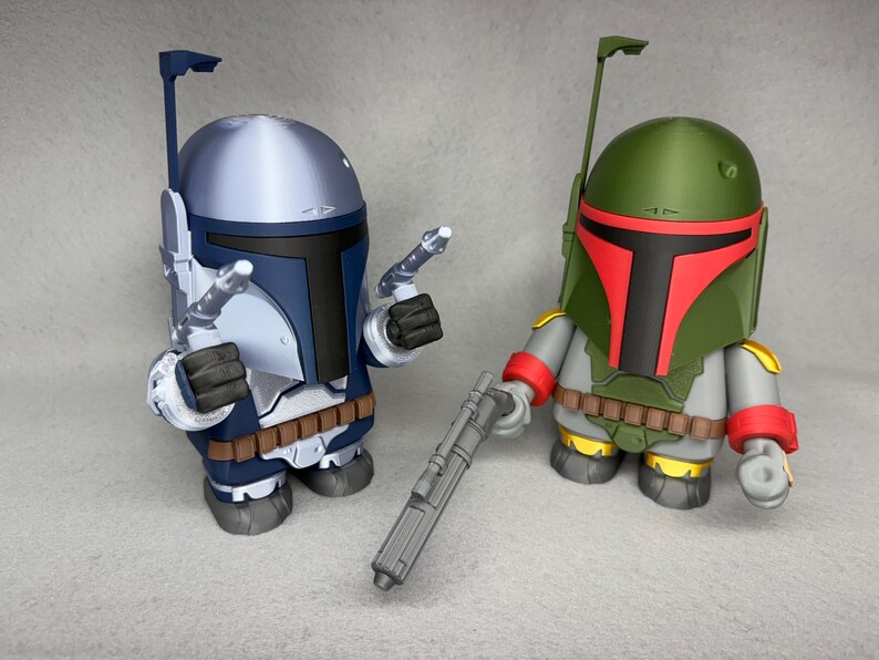 May include: Two Star Wars action figures, one in blue and silver and the other in green and red, stand on a gray surface. The blue and silver figure is holding a blaster rifle. The green and red figure is holding a blaster pistol.
