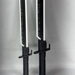 May include: Two black and white toy swords with a futuristic design. The blades are white with black edges and etched patterns. The handles are black with a ribbed texture and crossguard.