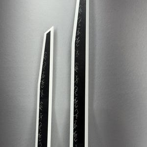 May include: Two tall, black and white abstract art pieces. The black sections feature white, stylized script. The pieces are set against a gray background. The shapes are long and angular, with a modern aesthetic.