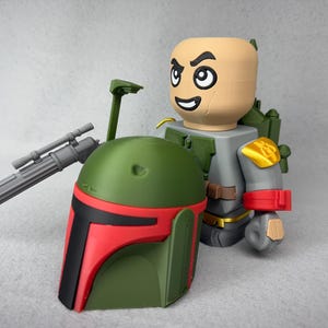 May include: A green and red Boba Fett helmet with a black visor. The helmet is next to a gray and gold action figure with a red and white arm band.
