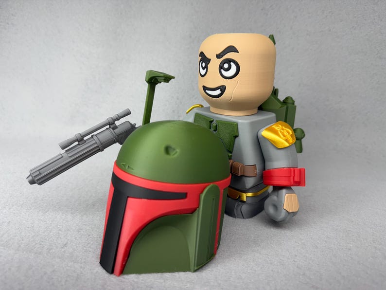 May include: A green and red Boba Fett helmet with a black visor. The helmet is next to a gray and gold action figure with a red and white arm band.