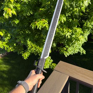 May include: A black and silver sword with a textured blade and a black handle. The sword is held against a backdrop of green foliage and a brown deck railing.