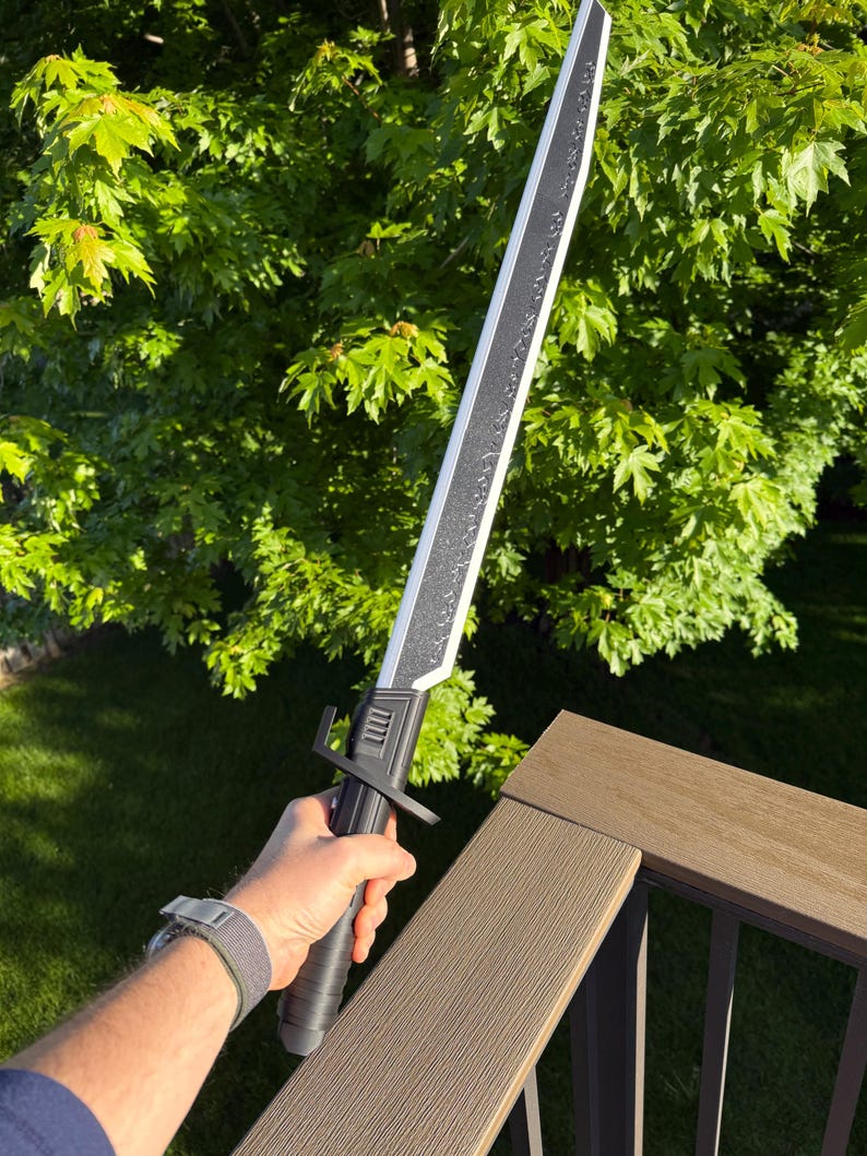 May include: A black and silver sword with a textured blade and a black handle. The sword is held against a backdrop of green foliage and a brown deck railing.