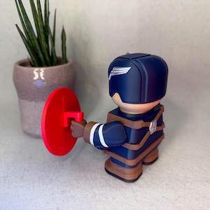 Op de afbeelding: Een marineblauwe superheldenfiguur met een rood schild. Het beeldje heeft een helm met een wit vleugelontwerp en bruine bandjes. Op de achtergrond staat een kleine plant in een pot.