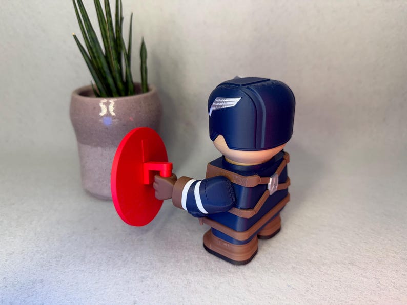 Op de afbeelding: Een marineblauwe superheldenfiguur met een rood schild. Het beeldje heeft een helm met een wit vleugelontwerp en bruine bandjes. Op de achtergrond staat een kleine plant in een pot.