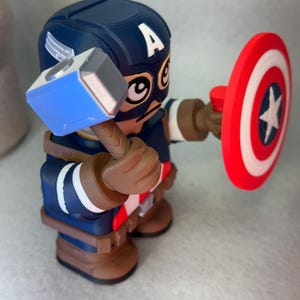 Op de afbeelding: Een 3D-geprinte Captain America-figuur, met een hamer en schild. De figuur is voornamelijk blauw, bruin en rood, met een witte ster op het schild. De helm heeft een witte "A".