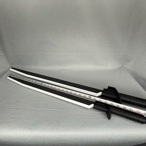 May include: Two black and white training swords with a ruler laid across them. The swords have a black handle and a white blade with a black stripe. The ruler is white with black markings.