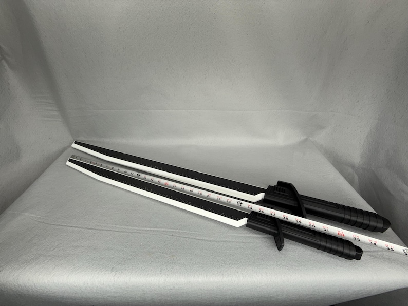 May include: Two black and white training swords with a ruler laid across them. The swords have a black handle and a white blade with a black stripe. The ruler is white with black markings.