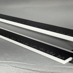 May include: Two black and white swords with decorative etchings along the black blade. The swords have a white edge and a black handle. The blades are angled towards the viewer.