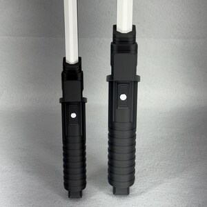 May include: Two black toy lightsabers with white blades. The hilts are cylindrical with ribbed and button details. One hilt is slightly larger than the other. The white blades are rectangular and extend from the top of the hilts.