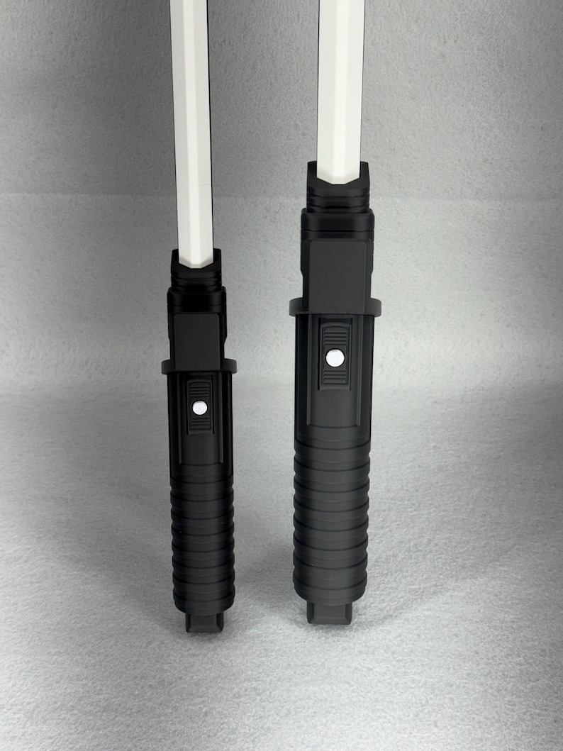 May include: Two black toy lightsabers with white blades. The hilts are cylindrical with ribbed and button details. One hilt is slightly larger than the other. The white blades are rectangular and extend from the top of the hilts.