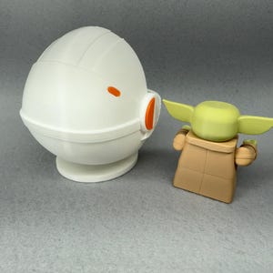 May include: A white, round, dome-shaped toy with an orange stripe and a small orange detail on the side.  The toy is next to a small, green, and brown toy with large ears.