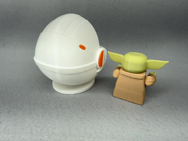 May include: A white, round, dome-shaped toy with an orange stripe and a small orange detail on the side.  The toy is next to a small, green, and brown toy with large ears.