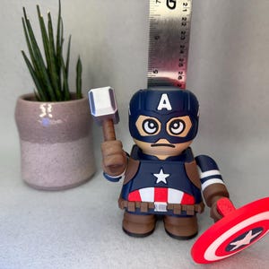 Op de afbeelding: Een Captain America-beeldje met een hamer en schild. Het beeldje is voornamelijk blauw, rood en wit, met een ster op de borst en het schild. Een kleine plantenpot staat op de achtergrond. Het beeldje is ongeveer 13 cm hoog.