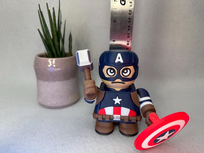 Op de afbeelding: Een Captain America-beeldje met een hamer en schild. Het beeldje is voornamelijk blauw, rood en wit, met een ster op de borst en het schild. Een kleine plantenpot staat op de achtergrond. Het beeldje is ongeveer 13 cm hoog.