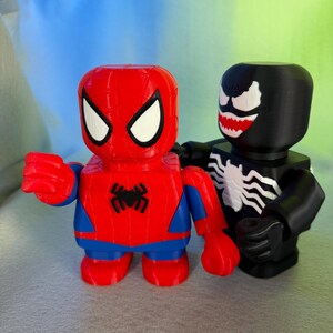 May include: A red and blue Spider-Man figurine stands next to a black Venom figurine. Both figurines are in a blocky style, with simple features and a cartoonish appearance.