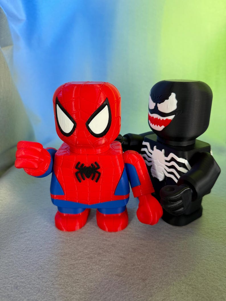 May include: A red and blue Spider-Man figurine stands next to a black Venom figurine. Both figurines are in a blocky style, with simple features and a cartoonish appearance.