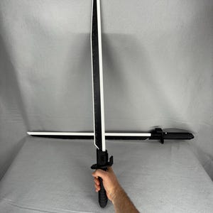 May include: Two black and white toy swords. The swords have a black handle and a white blade with a black stripe down the center. One sword is held by a person, and the other is lying horizontally.
