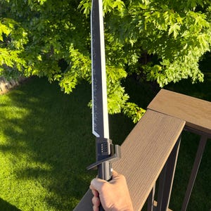 May include: A black and white sword with a long blade and a detailed handle. The blade has a white edge and a black center with decorative markings. The handle is black with a textured grip. The sword is held against a background of green foliage.