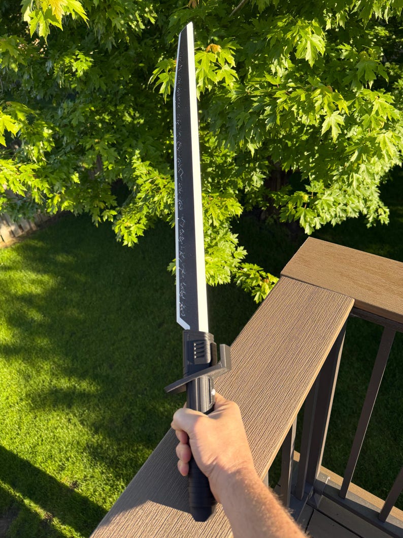 May include: A black and white sword with a long blade and a detailed handle. The blade has a white edge and a black center with decorative markings. The handle is black with a textured grip. The sword is held against a background of green foliage.