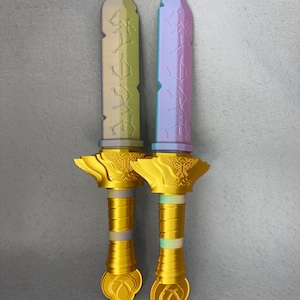 May include: Two decorative toy swords. One is pale green, the other is purple and blue. Both have gold handles and blades with etched designs. The handles have a detailed, ornate design. The swords are displayed against a gray background.
