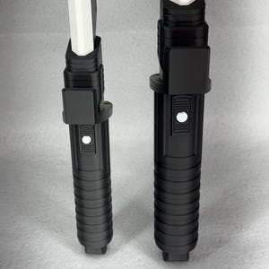 May include: Two black lightsaber hilts with white blades. The hilts have a ribbed design and a button. The blades are rectangular and white. The lightsabers are standing upright against a light gray background.