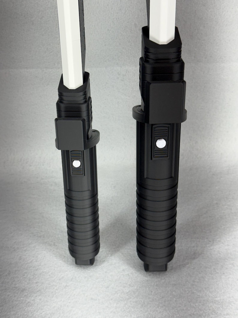 May include: Two black lightsaber hilts with white blades. The hilts have a ribbed design and a button. The blades are rectangular and white. The lightsabers are standing upright against a light gray background.
