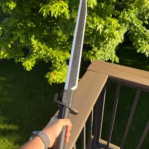 May include: A black and silver sword with a textured handle and a straight blade. The sword is held against a backdrop of green foliage and a brown deck railing. The blade has a decorative design along its edge.