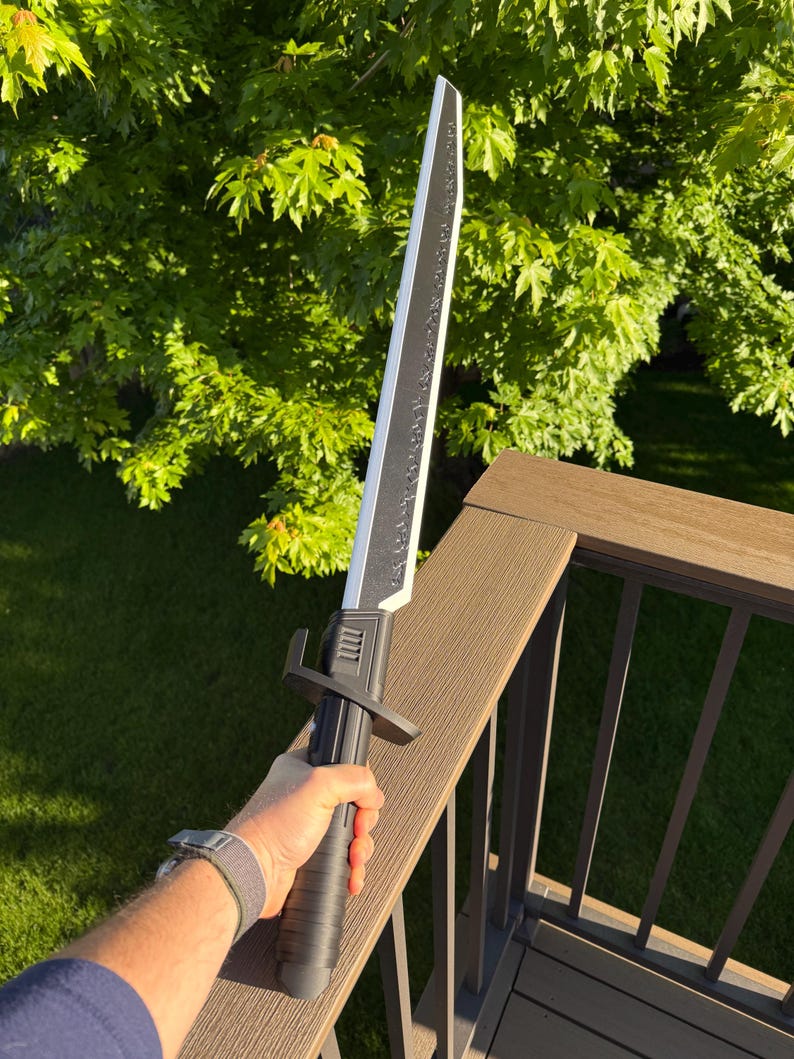 May include: A black and silver sword with a textured handle and a straight blade. The sword is held against a backdrop of green foliage and a brown deck railing. The blade has a decorative design along its edge.