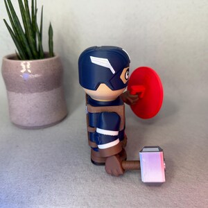 Op de afbeelding: Een Captain America-beeldje met een blauwe helm en schild, met een hamer. Het beeldje draagt een blauw en bruin uniform. Een kleine plant in een pot staat op de achtergrond.